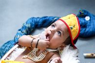Cute baby dressed in traditional Indian clothing lying down, wearing jewelry and a vibrant hat.