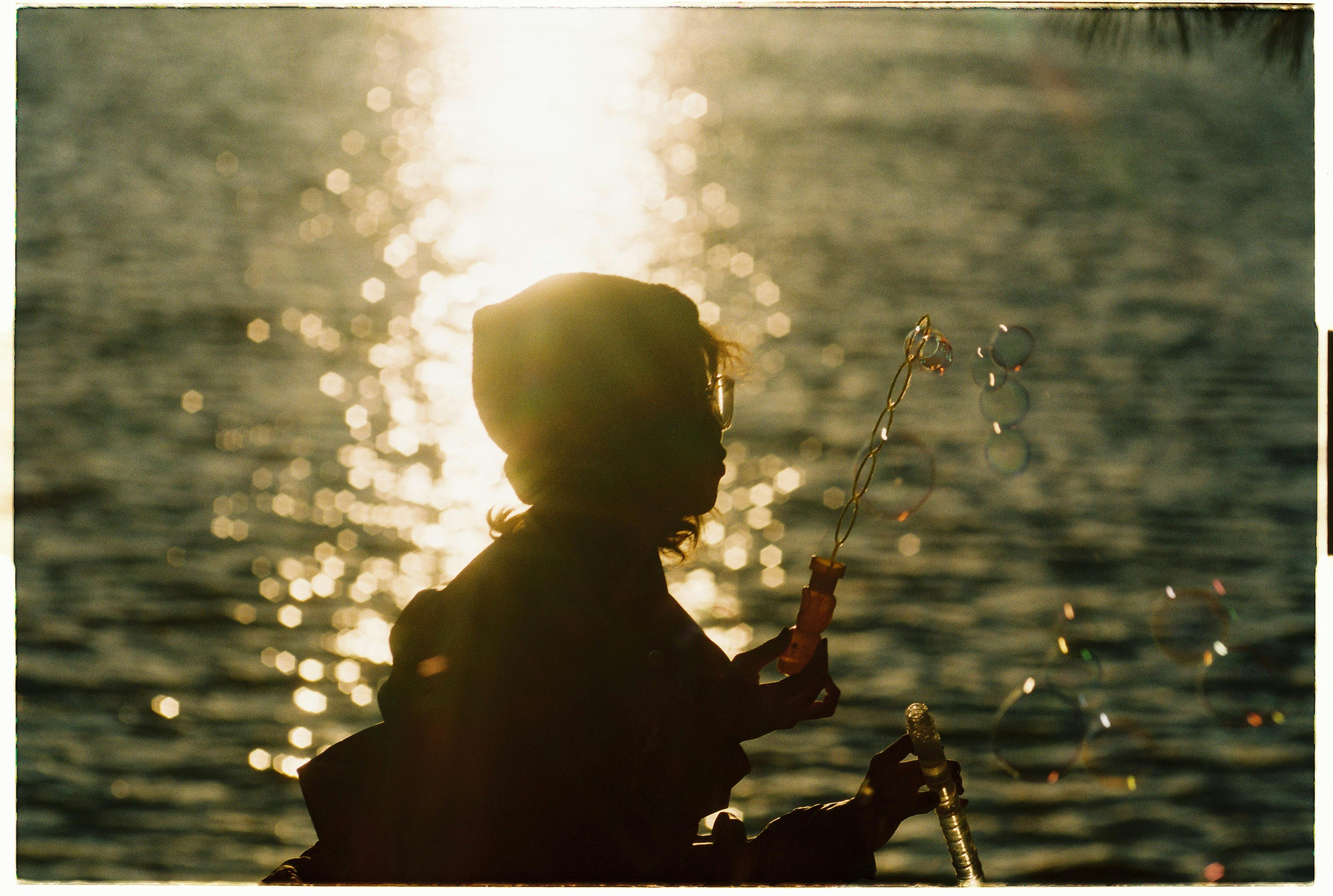 A child blowing bubbles with a sunset reflecting off the water, creating a serene silhouette effect.