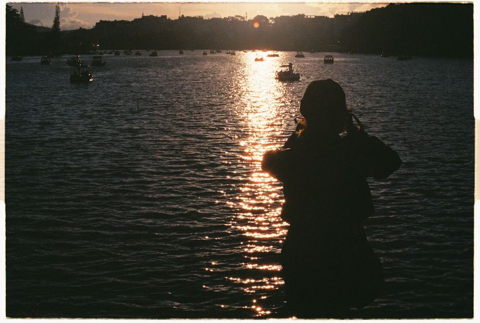 Silhouette of a person by a calm body of water with boats at sunset.