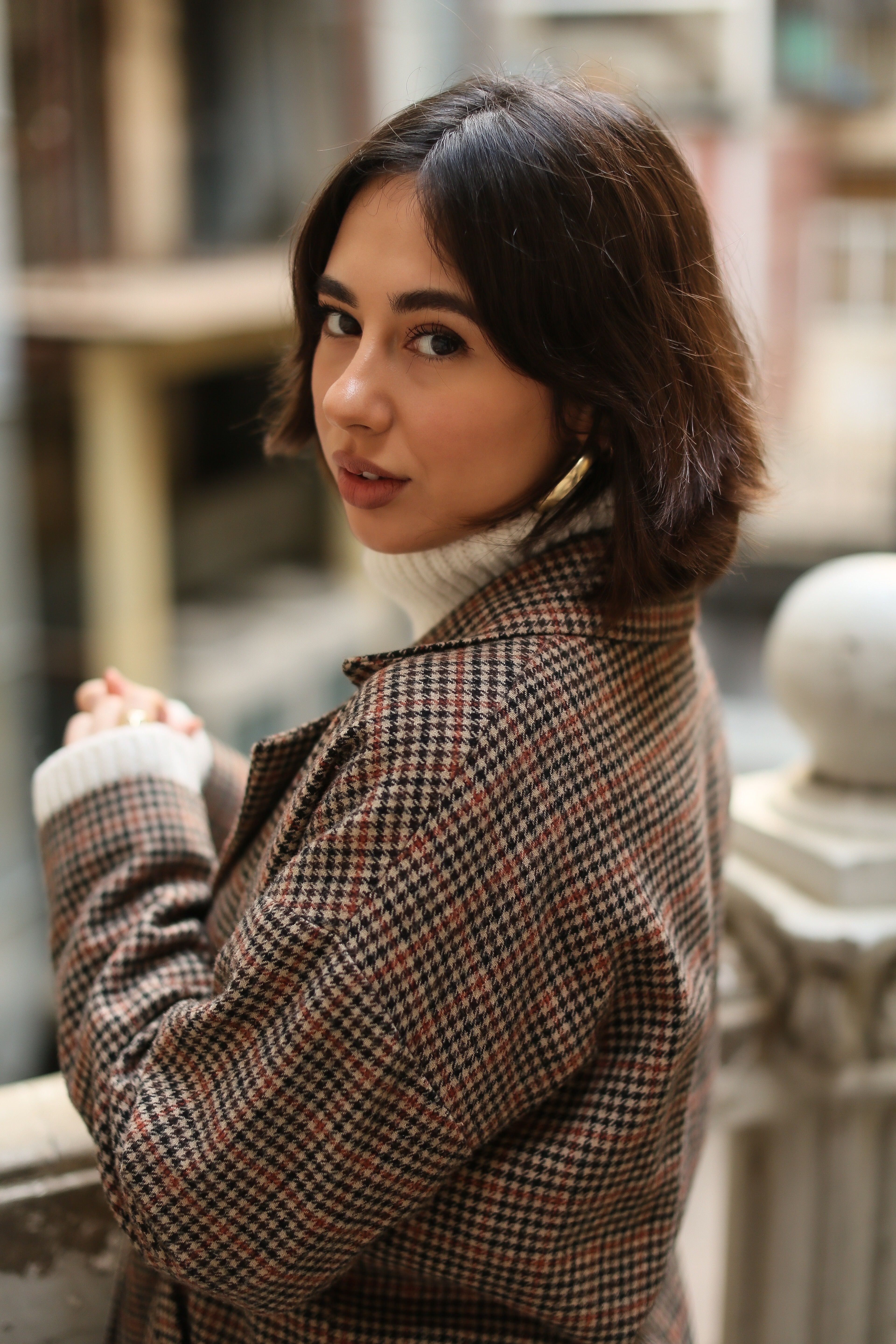 Young woman in plaid coat poses outdoors, showcasing modern fashion style in urban environment.