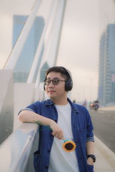 Young man with headphones holding a sunflower on a bridge in urban Vietnam cityscape.