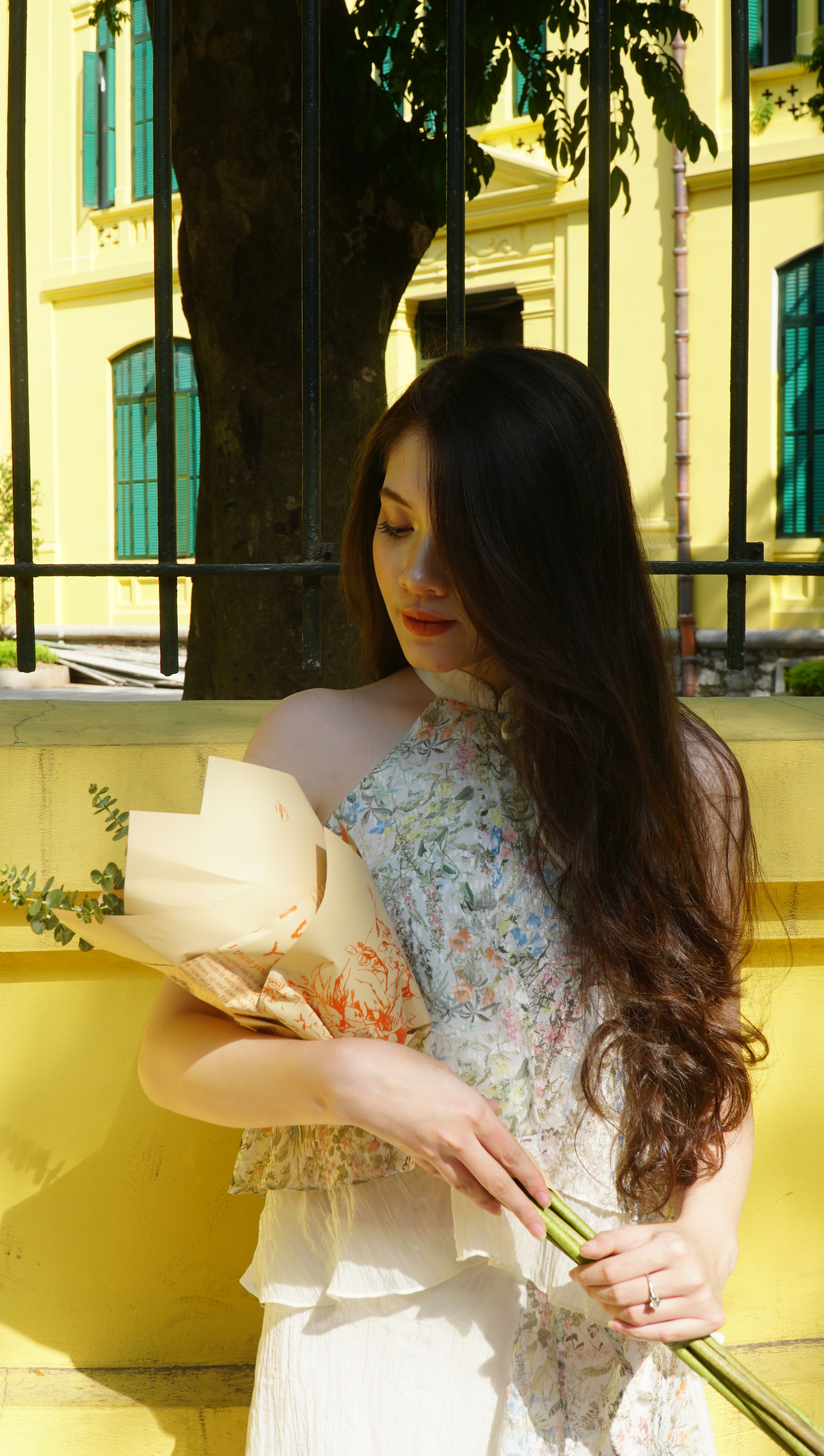 A woman in a floral dress holds a bouquet against a yellow urban backdrop in daylight.