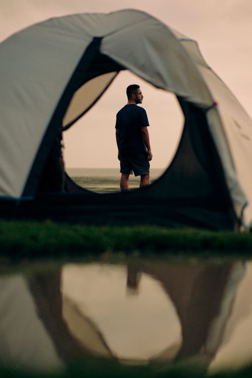 Free A man stands next to a tent by a reflective water surface at sunset, creating a serene outdoor scene. Stock Photo