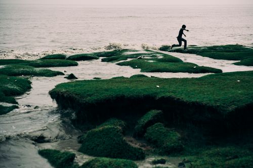 Free Silhouette of a child joyfully leaping across mossy coastal rocks near the sea during daylight. Stock Photo