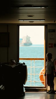 A child stands on a ship, gazing at the sea through an open door, with a cargo vessel in the distance.