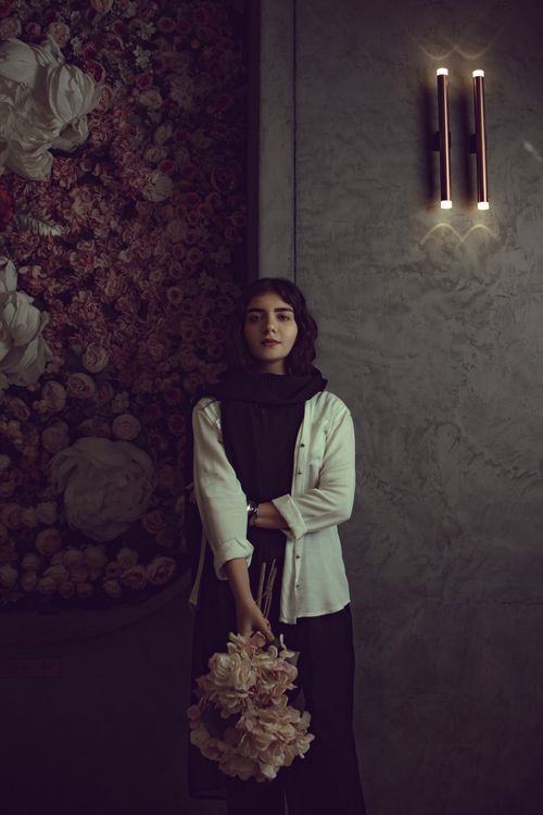 Free Woman in white cardigan holding flowers stands against floral wall in dimly lit room. Stock Photo