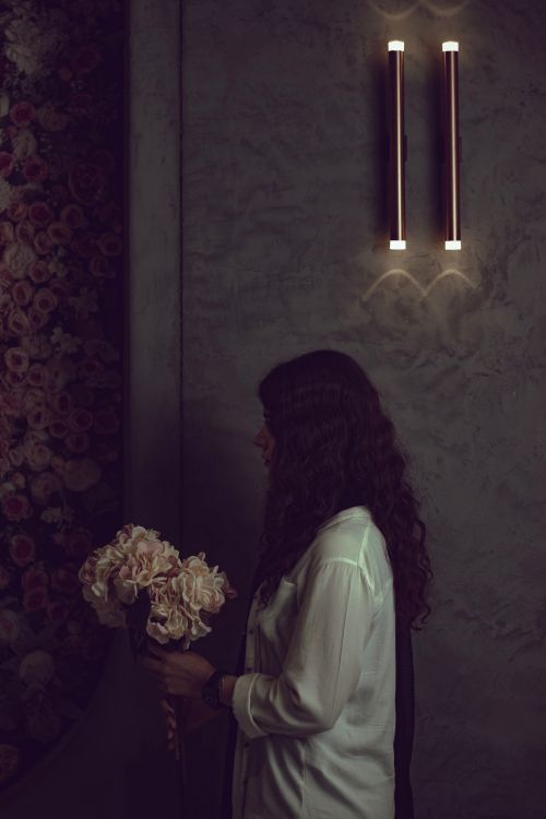 Free A woman with wavy hair holds a bouquet of flowers against a dimly lit, textured wall indoors. Stock Photo