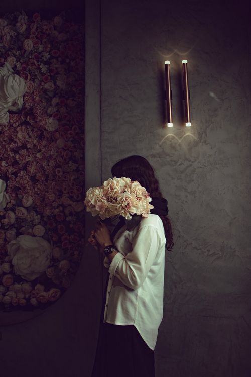 Free A woman holds a bouquet of flowers in front of her face, standing by a textured wall with dim lighting. Stock Photo