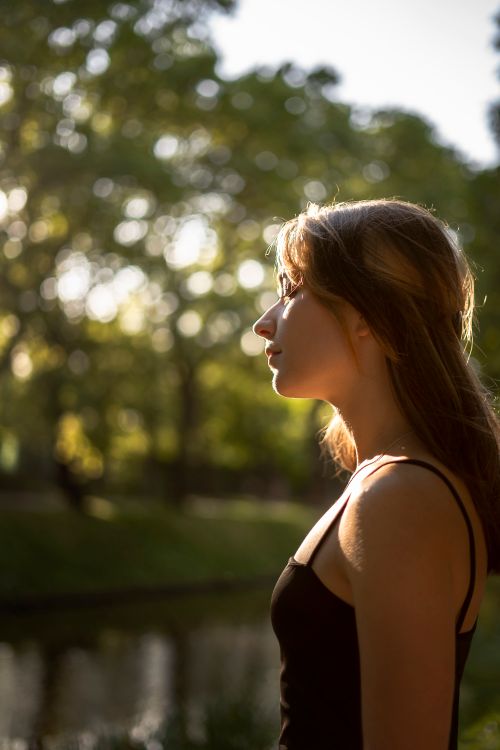Free Side profile of a woman with long brown hair standing outdoors in a sunlit park. Stock Photo