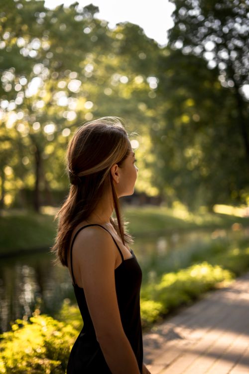 Free A young woman in a black dress stands by a tree-lined path during sunset. Perfect for lifestyle themes. Stock Photo