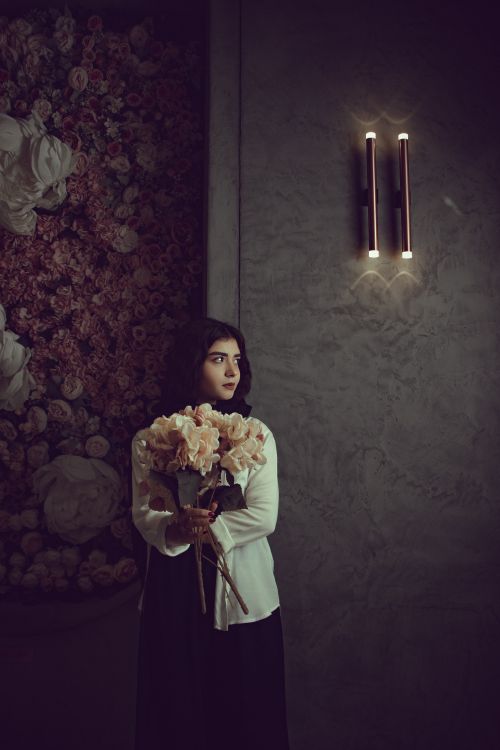 Free Moody portrait of a woman holding a bouquet, standing by a decorated wall and sconce light. Stock Photo