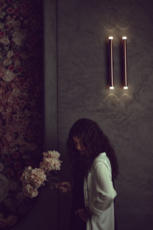 Free A young woman in a moody room with flowers and wall lamp. Stock Photo