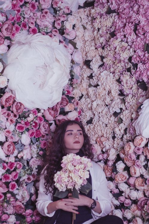 Free A young woman with wavy hair poses against a lush floral wall, holding a bouquet. Stock Photo