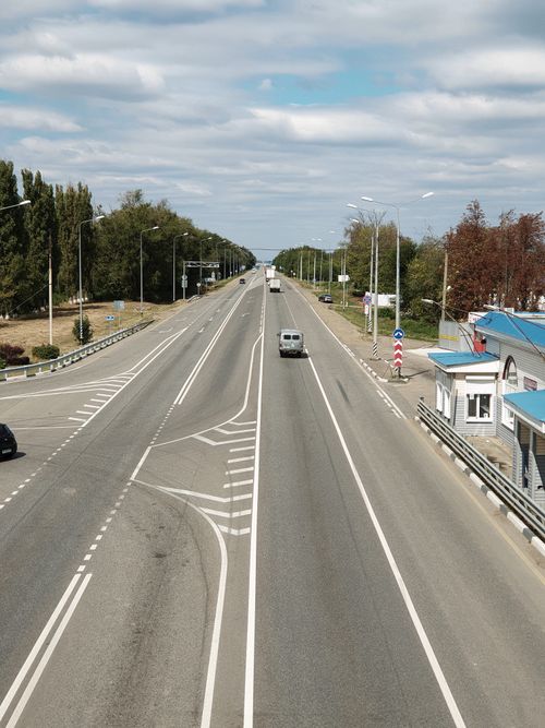 Free A drone captures an aerial shot of a wide, empty rural road with sparse vehicles surrounded by trees. Stock Photo