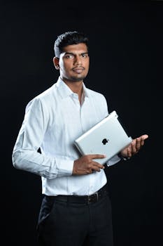 Portrait of a confident man holding a tablet against a black background, wearing a white shirt.