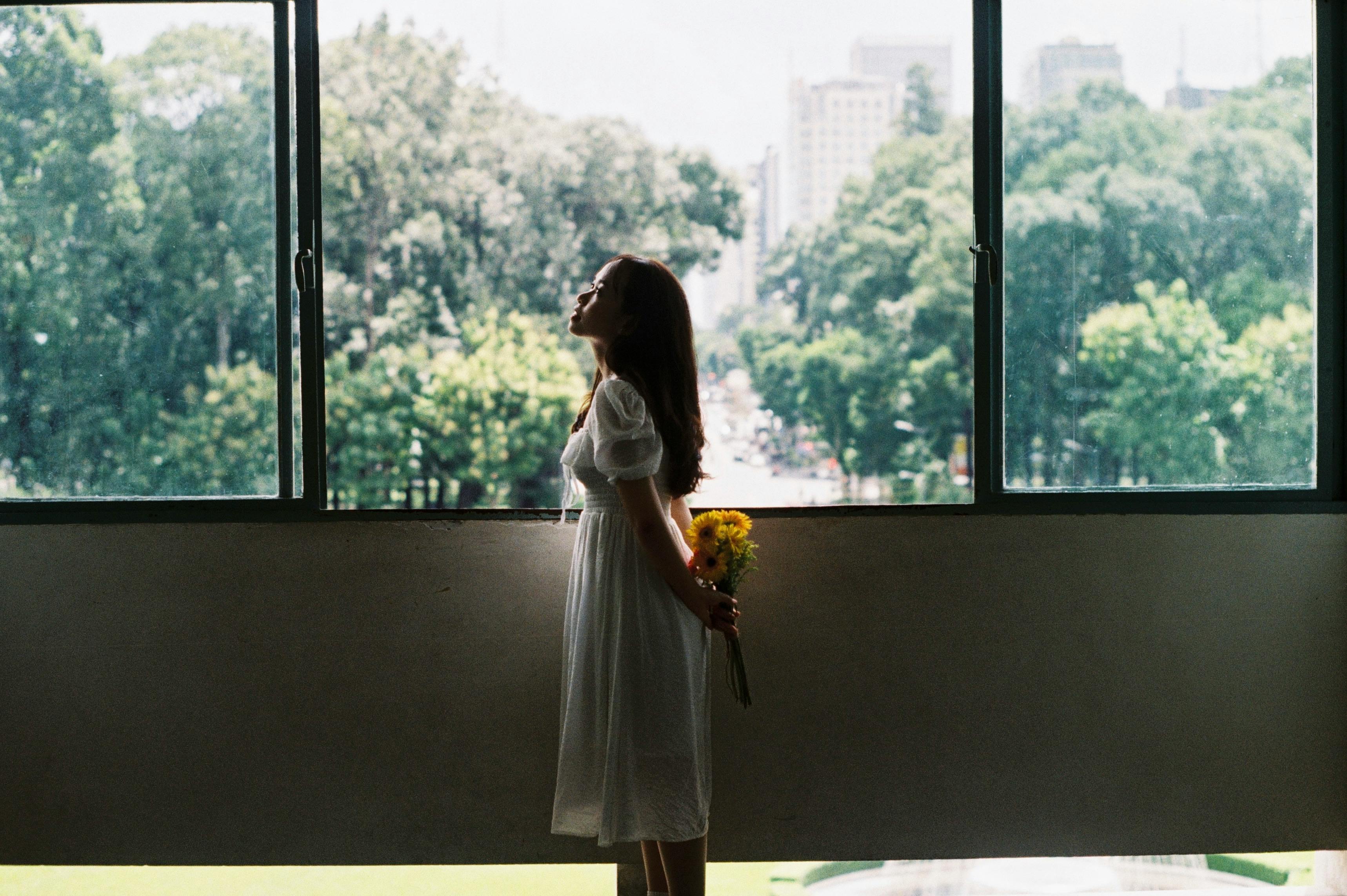 A serene moment of a woman in a white dress holding flowers indoors, embracing sunlight by a window.