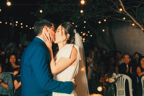 Free Newlywed couple sharing a romantic kiss under enchanting outdoor string lights. Stock Photo