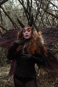 Dramatic fantasy costume portrait of a horned, winged woman standing in a forest.