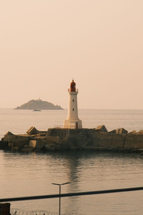 Free A tranquil view of a lighthouse on the rocky shore against a calm sea and distant island. Stock Photo