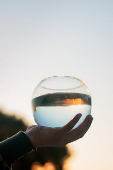 A serene image of a hand holding a glass fish bowl filled with water against a sunset background.