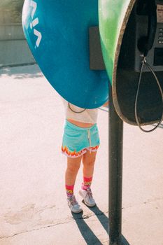 A child standing at a retro payphone booth on a sunny street in São José do Rio Preto, Brazil.
