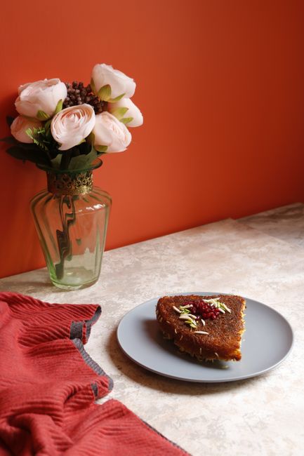 A beautifully plated dessert slice accompanied by a vase of flowers on a table with a red background.
