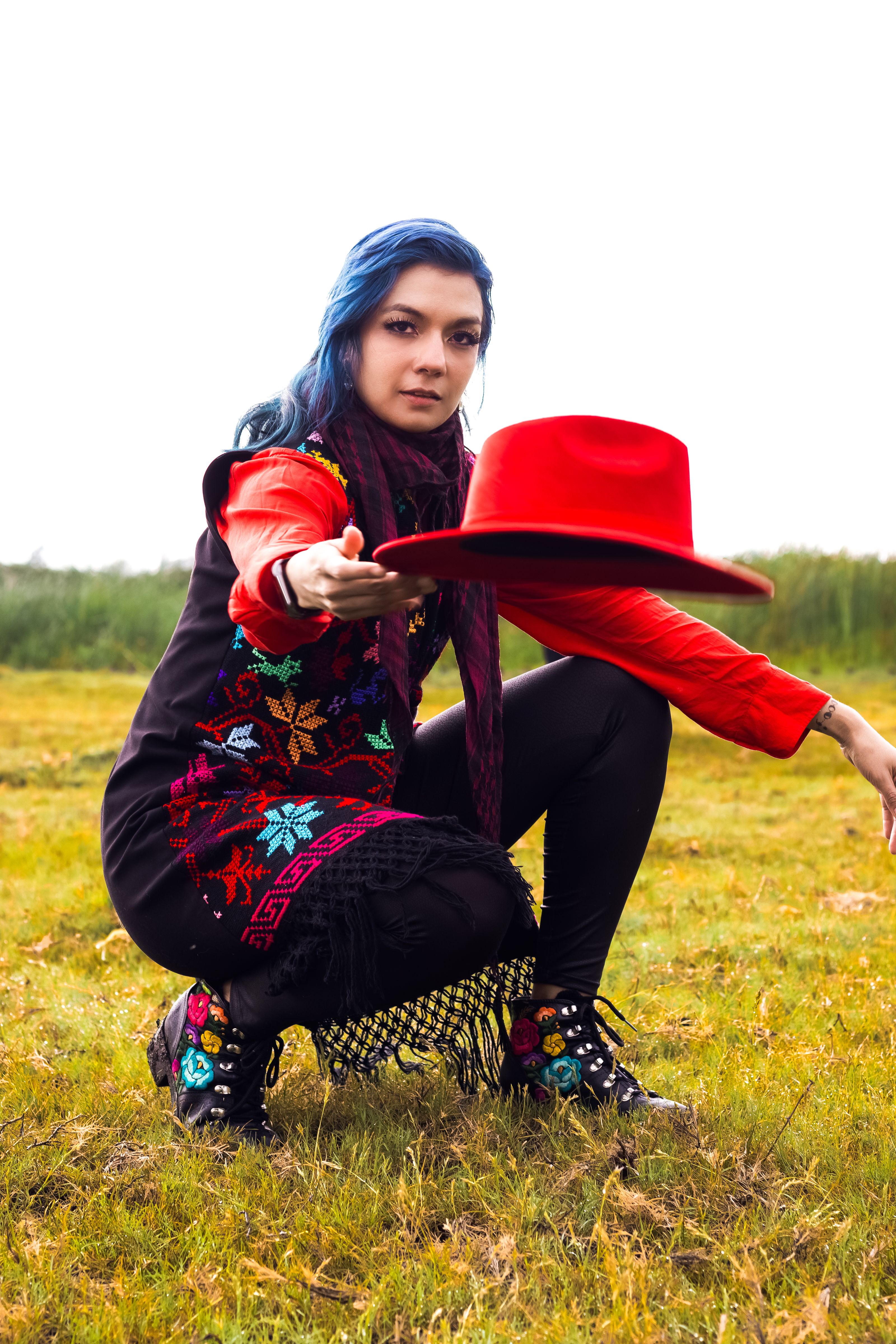 Mulher em traje tradicional brilhante posando com um chapéu vermelho em um campo gramado.