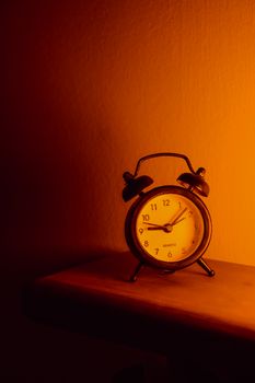 Rustic analog alarm clock casting a shadow on a shelf with warm orange lighting.