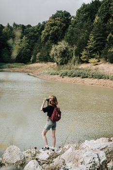A woman with a backpack takes photos by a tranquil lakeshore surrounded by a lush forest.