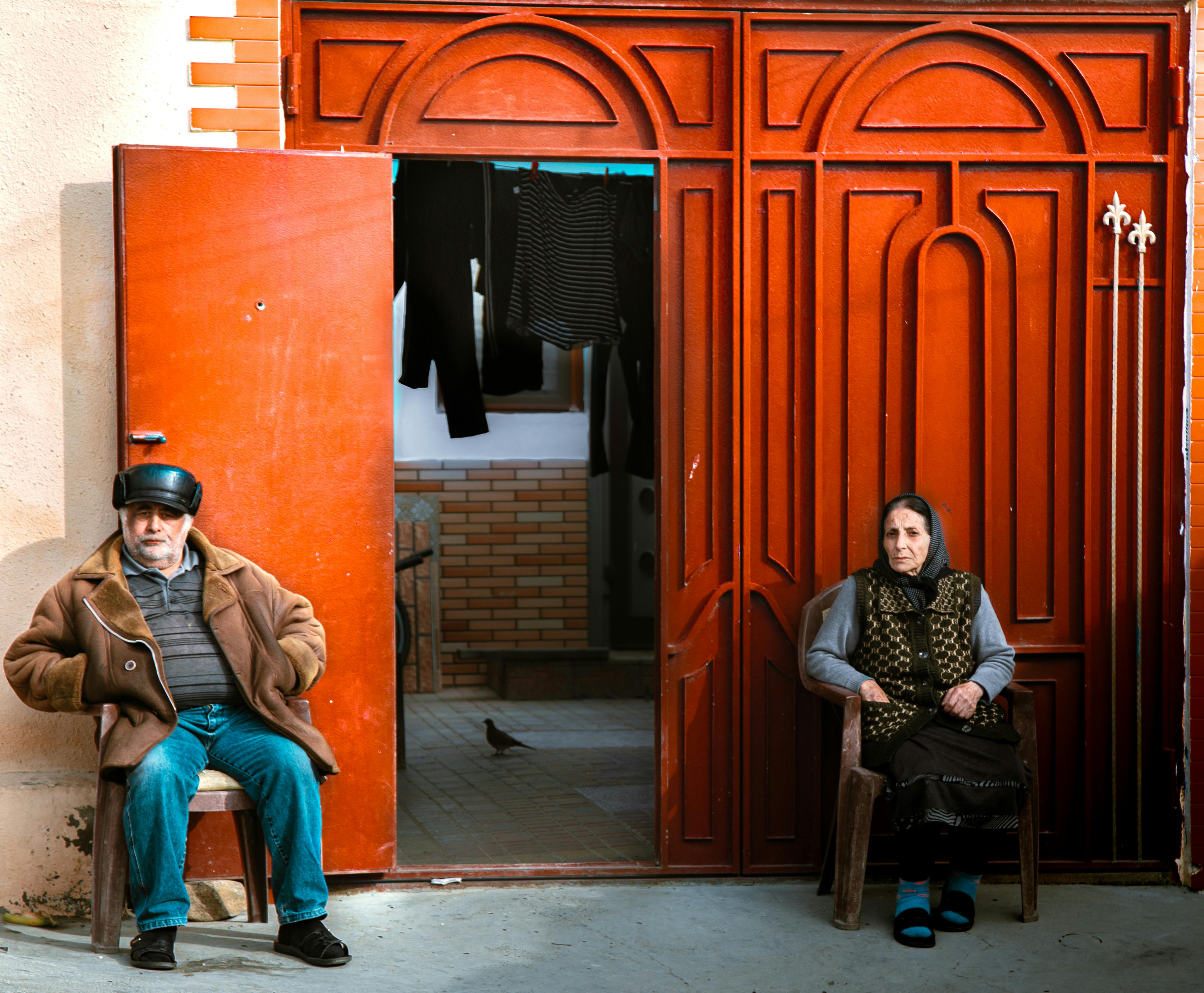 Serene elderly couple sitting by a red wooden doorway, enjoying the sunlight.