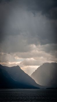 Moody mountain scene with rain clouds casting shadows over the sea.