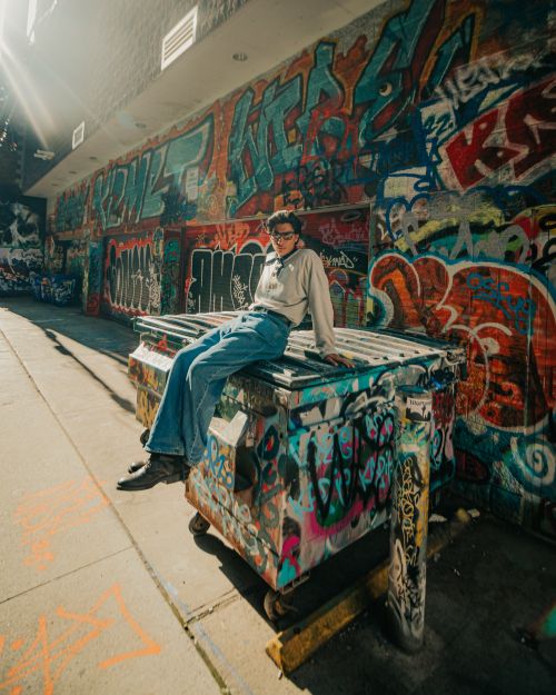Free A young man wearing eyeglasses sits on a graffiti-covered dumpster in a vibrant urban alley. Stock Photo