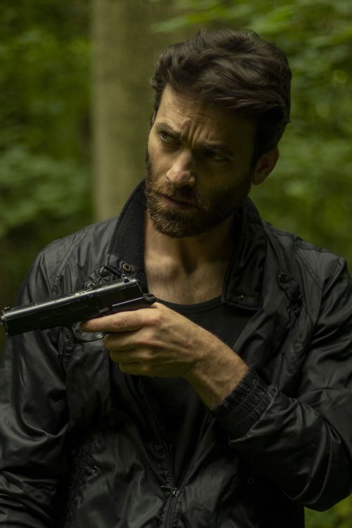 Free A man in a black jacket stands in a forest, holding a gun, portraying a tense, dramatic moment. Stock Photo