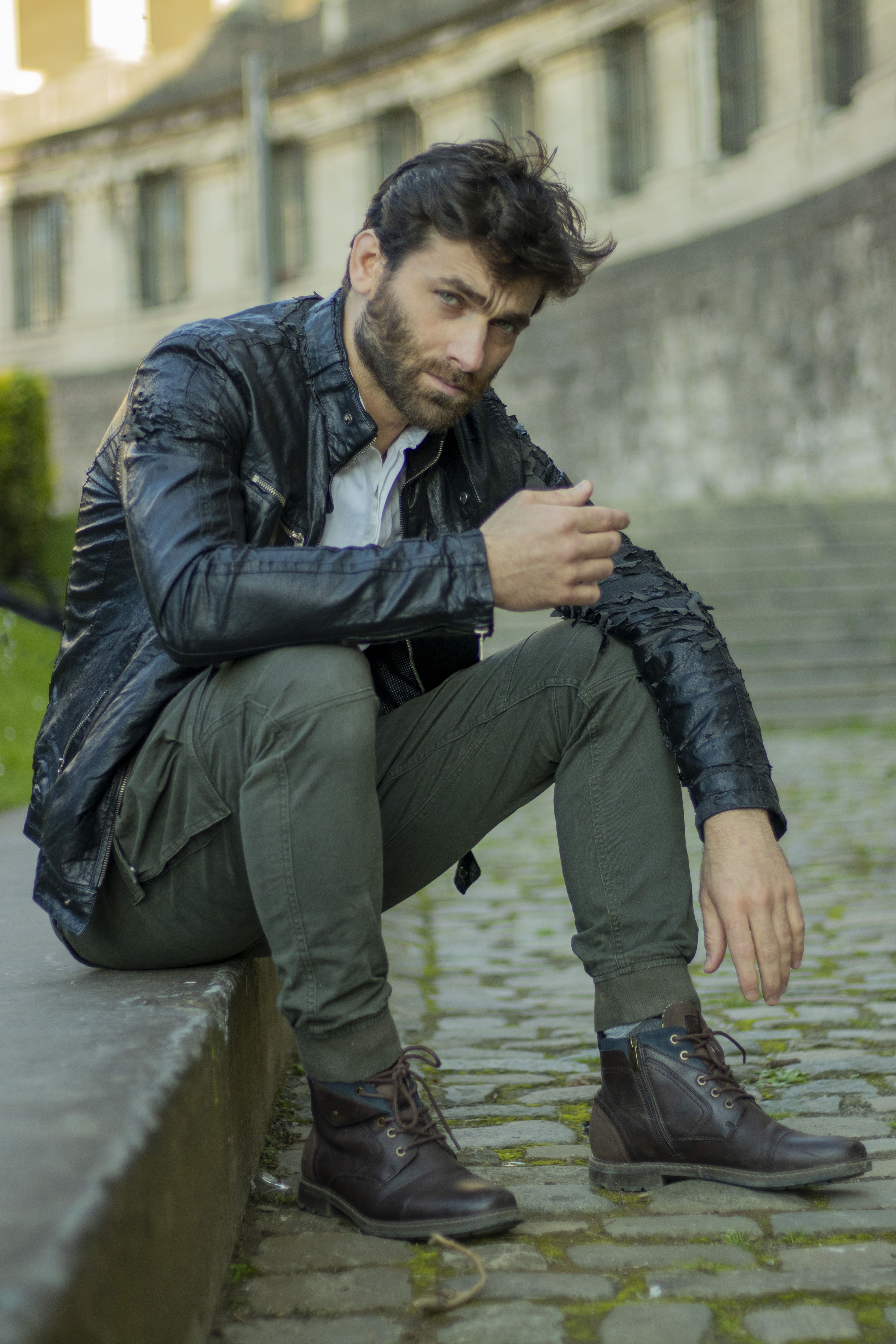 Fashionable man in a leather jacket sitting outdoors on a cobblestone path.