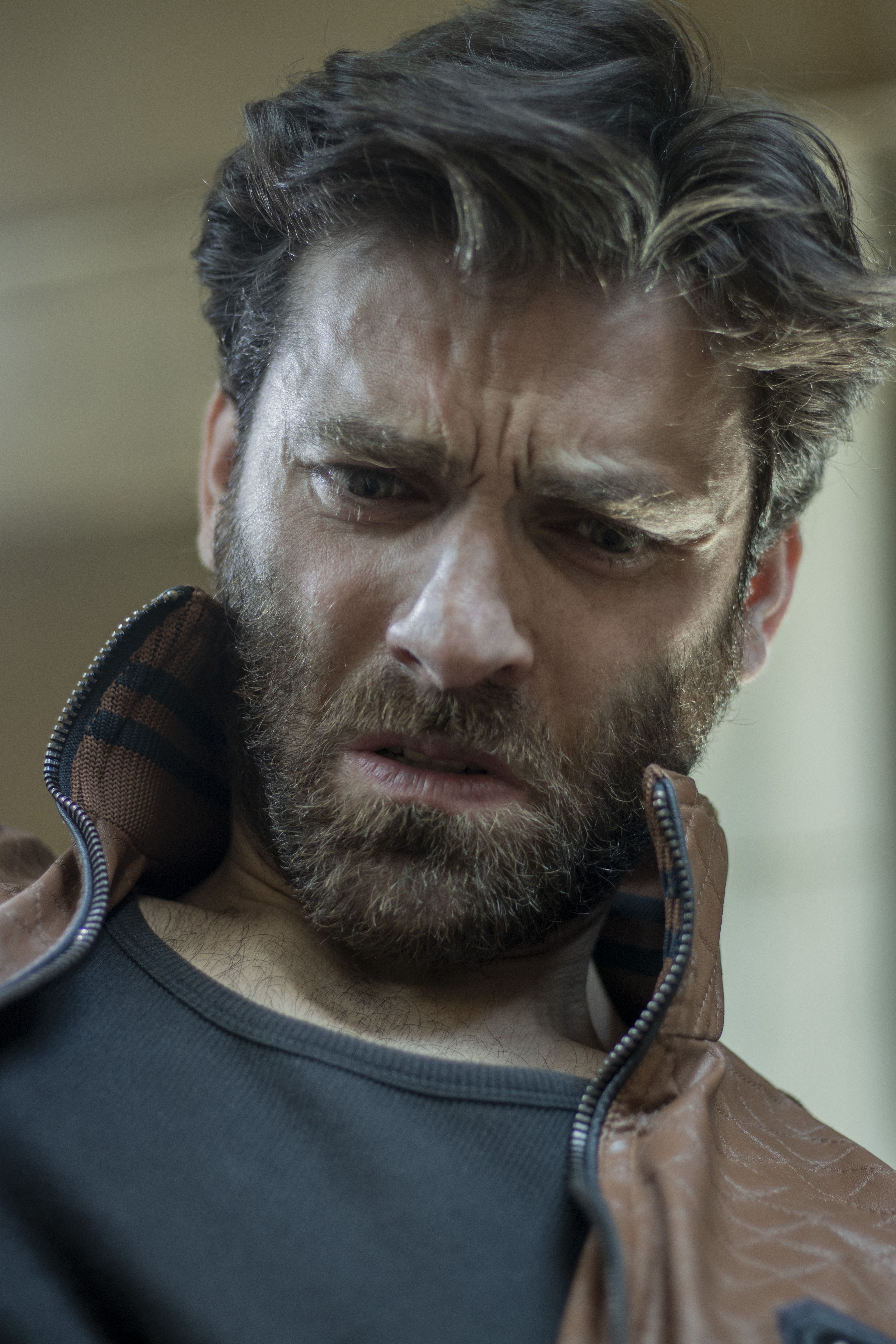 Close-up portrait of a bearded man looking down pensively, wearing a jacket indoors.