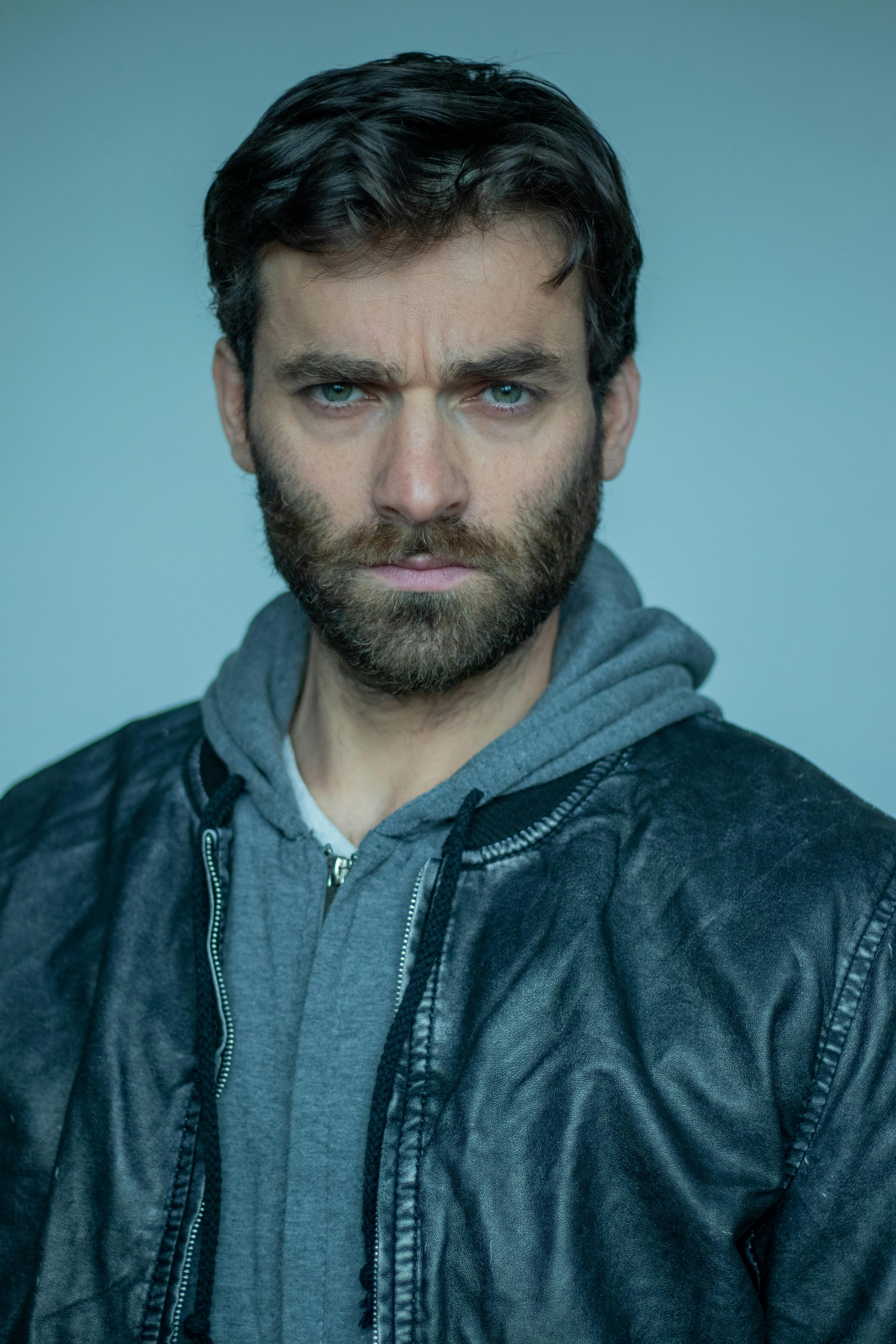 Portrait of a serious man with a beard, wearing a hoodie and jacket, captured in a studio setting.
