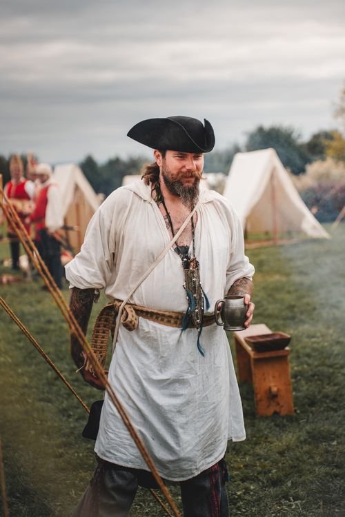 Free A male performer at a historical reenactment event, dressed in period costume, holding a cup outdoors. Stock Photo