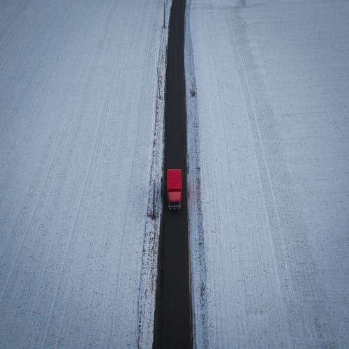 Drone view of red truck driving on straight roadway between snowy meadows in countryside on winter day