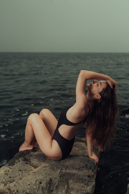 A woman in a black swimsuit sitting on a rock by the ocean