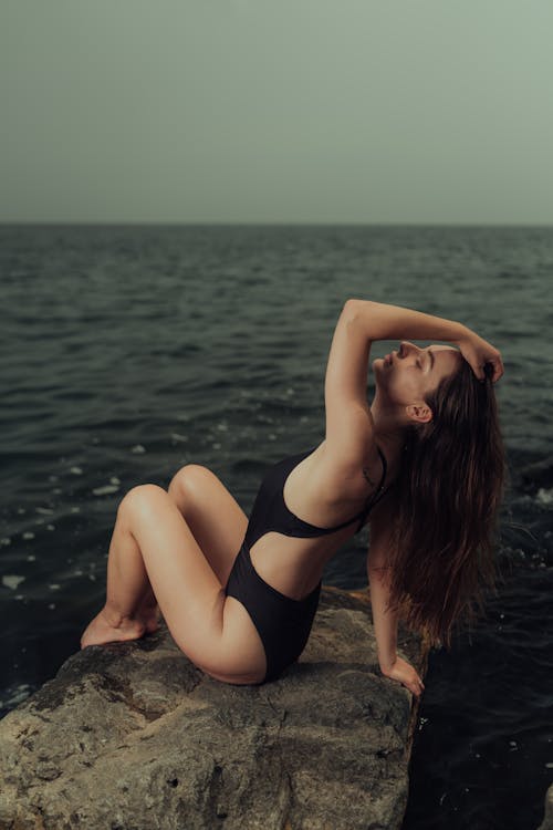 Free A woman in a black swimsuit sitting on a rock by the ocean Stock Photo