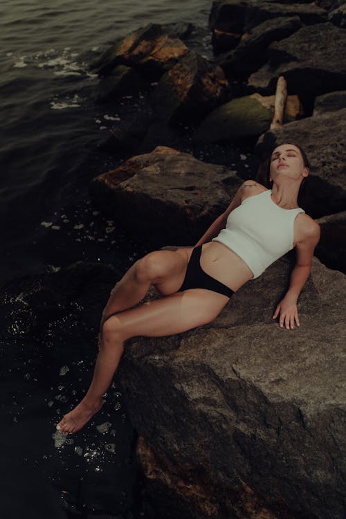 A woman in a white top and black bottoms is laying on rocks