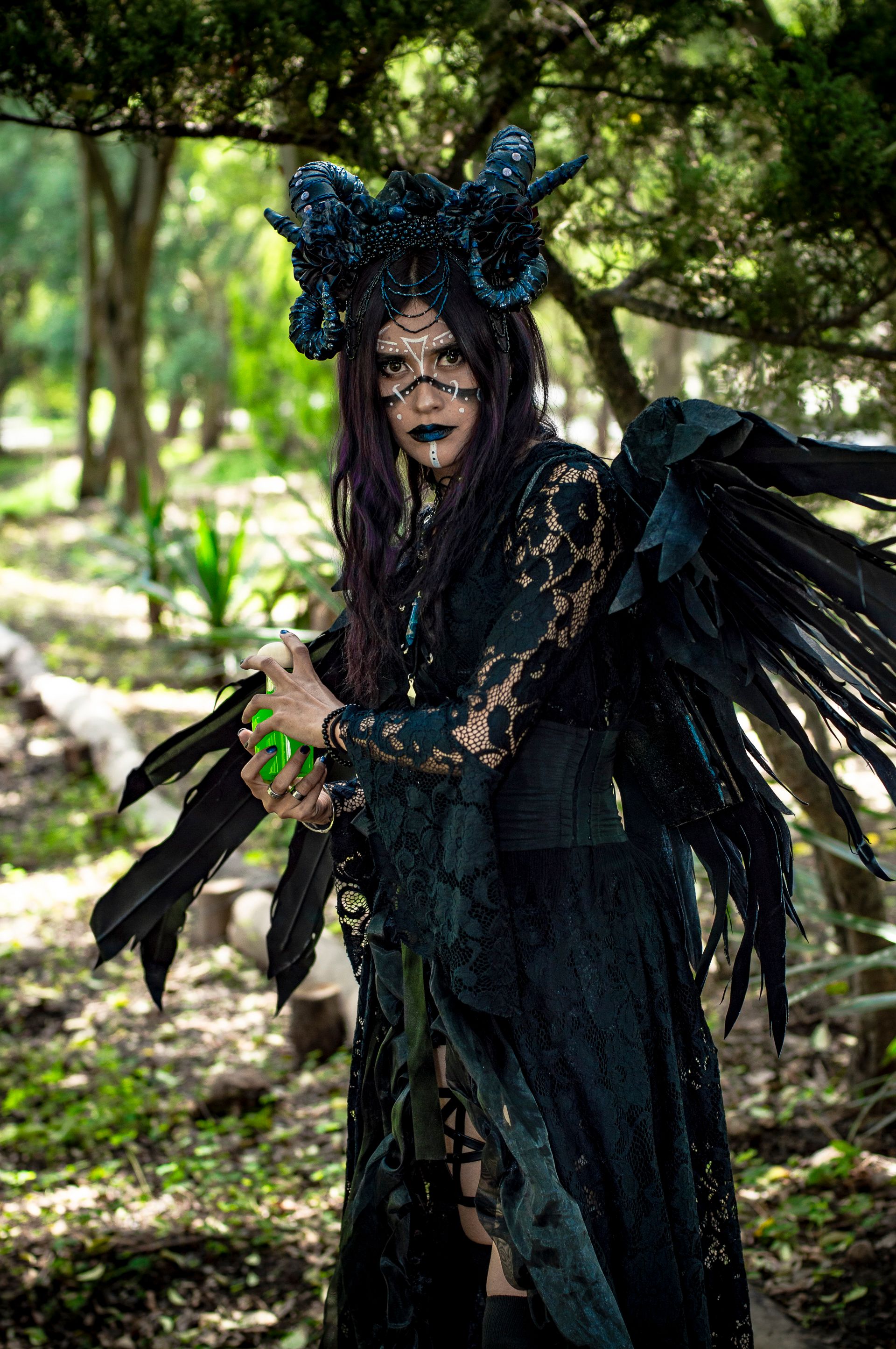 Mysterious woman in elaborate dark fantasy costume with wings holding a glowing elixir in a mystical forest setting.