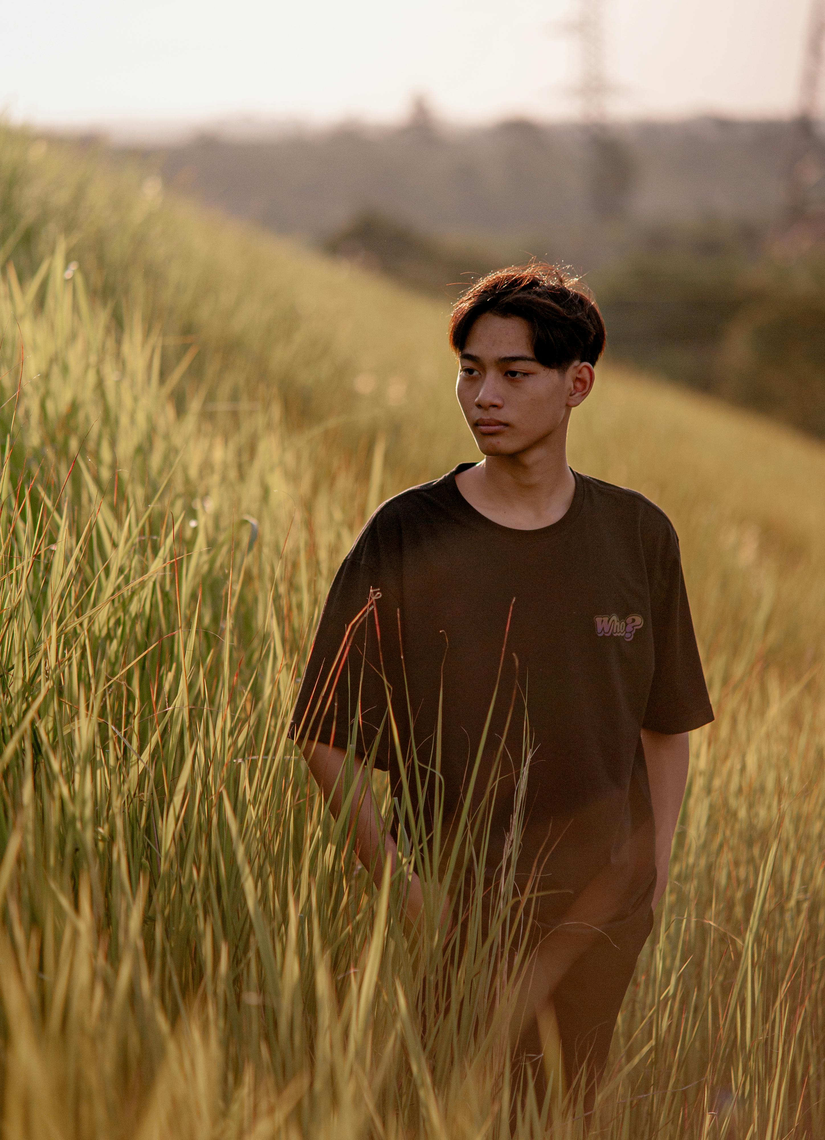 A young man in a black T-shirt standing amidst tall grass in a serene countryside landscape at sunset.