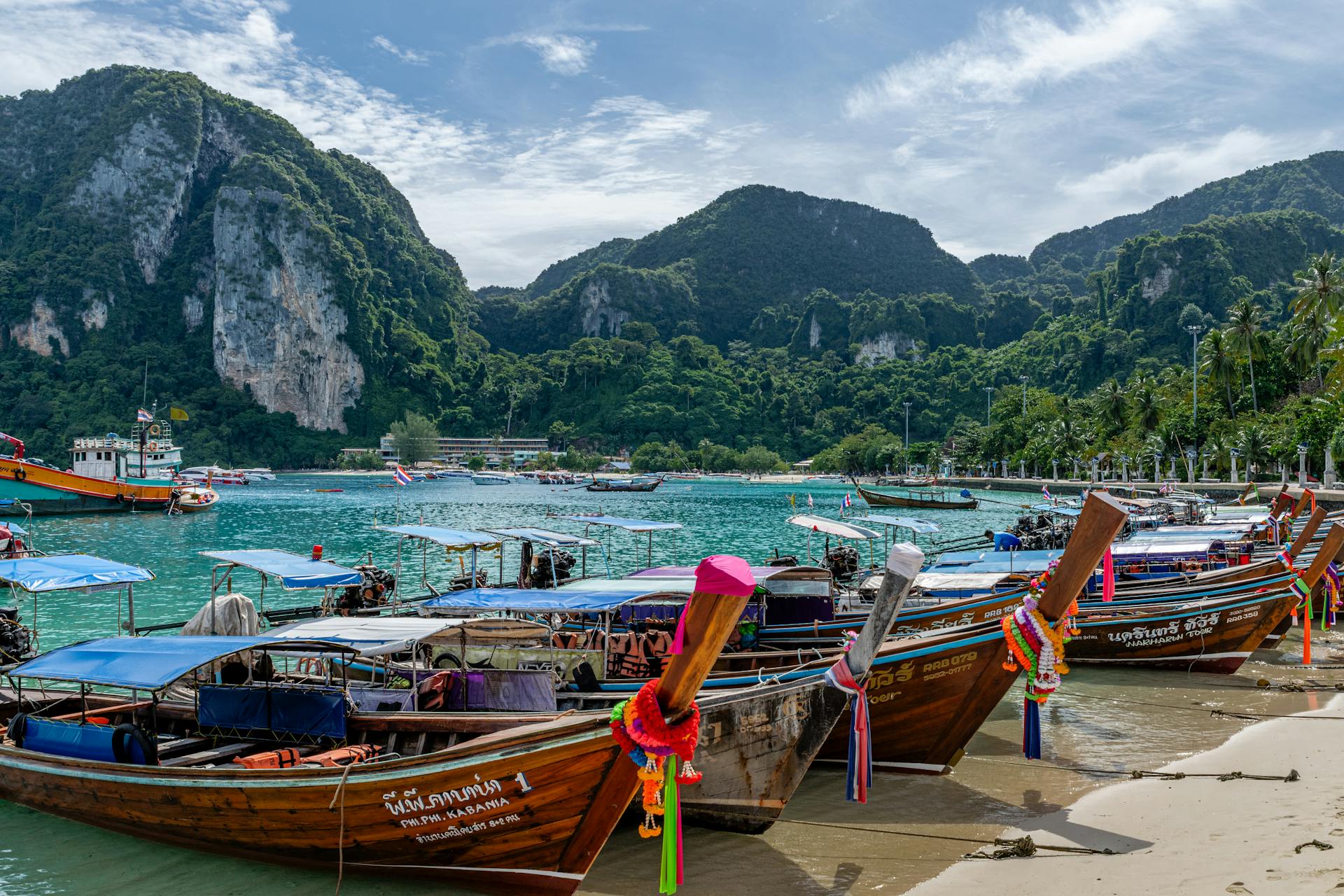 Longtail boats lined up at a stunning beach in Phuket, Thailand, with lush green mountains.