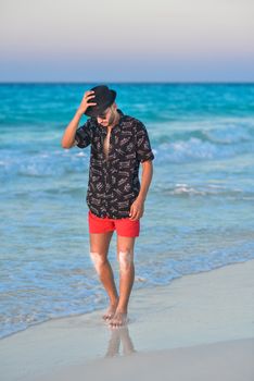 Man in black fedora and red shorts walking along a sunny tropical beach.