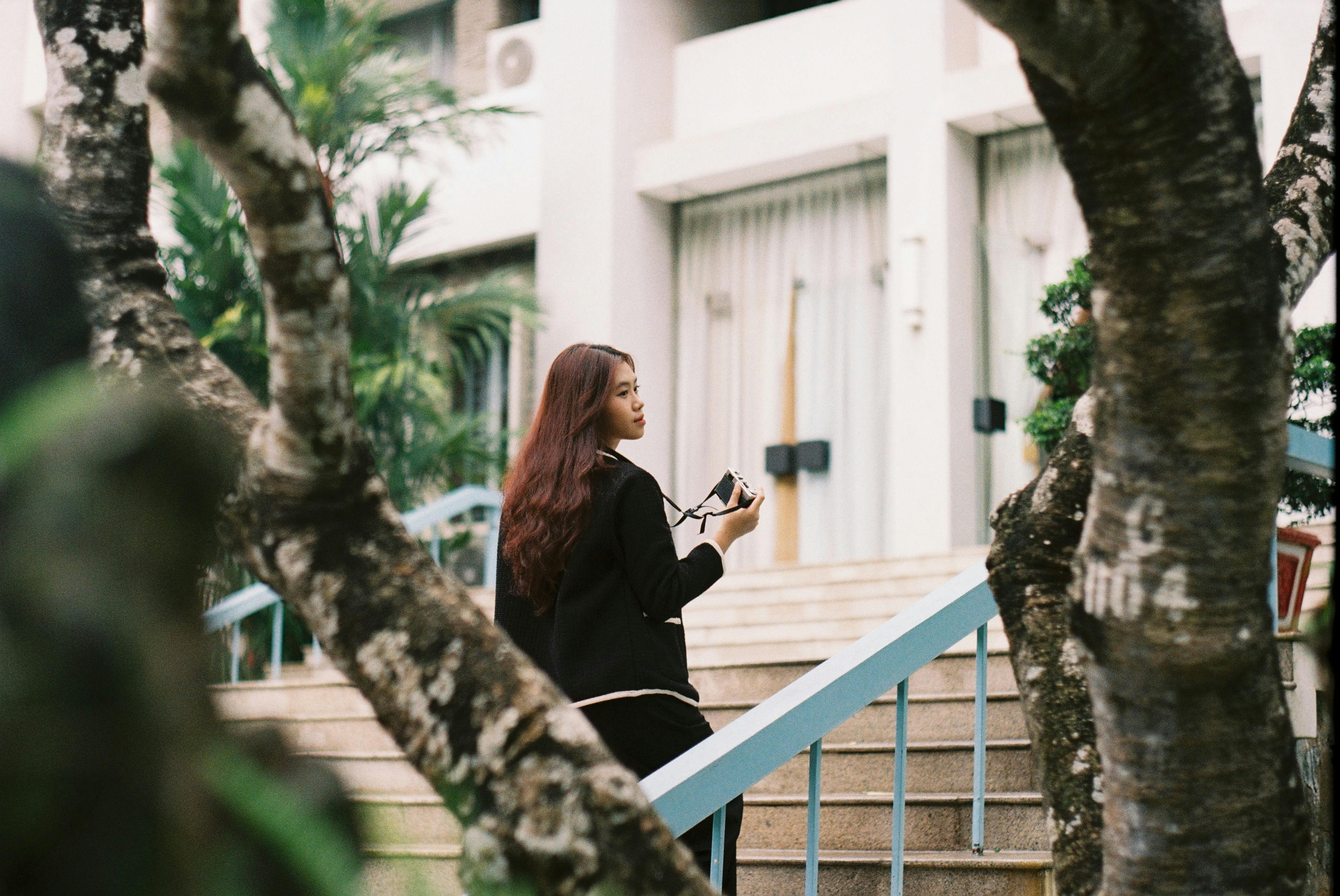 Free Back view of an Asian woman holding a camera, standing on steps in urban setting. Stock Photo