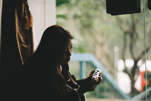 Young woman holding a camera, gazing out a rainy window with a thoughtful expression.