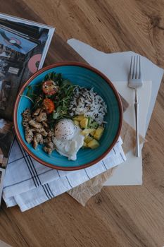 Top view of a colorful rice bowl with grilled chicken, vegetables, and poached egg on a wooden table.