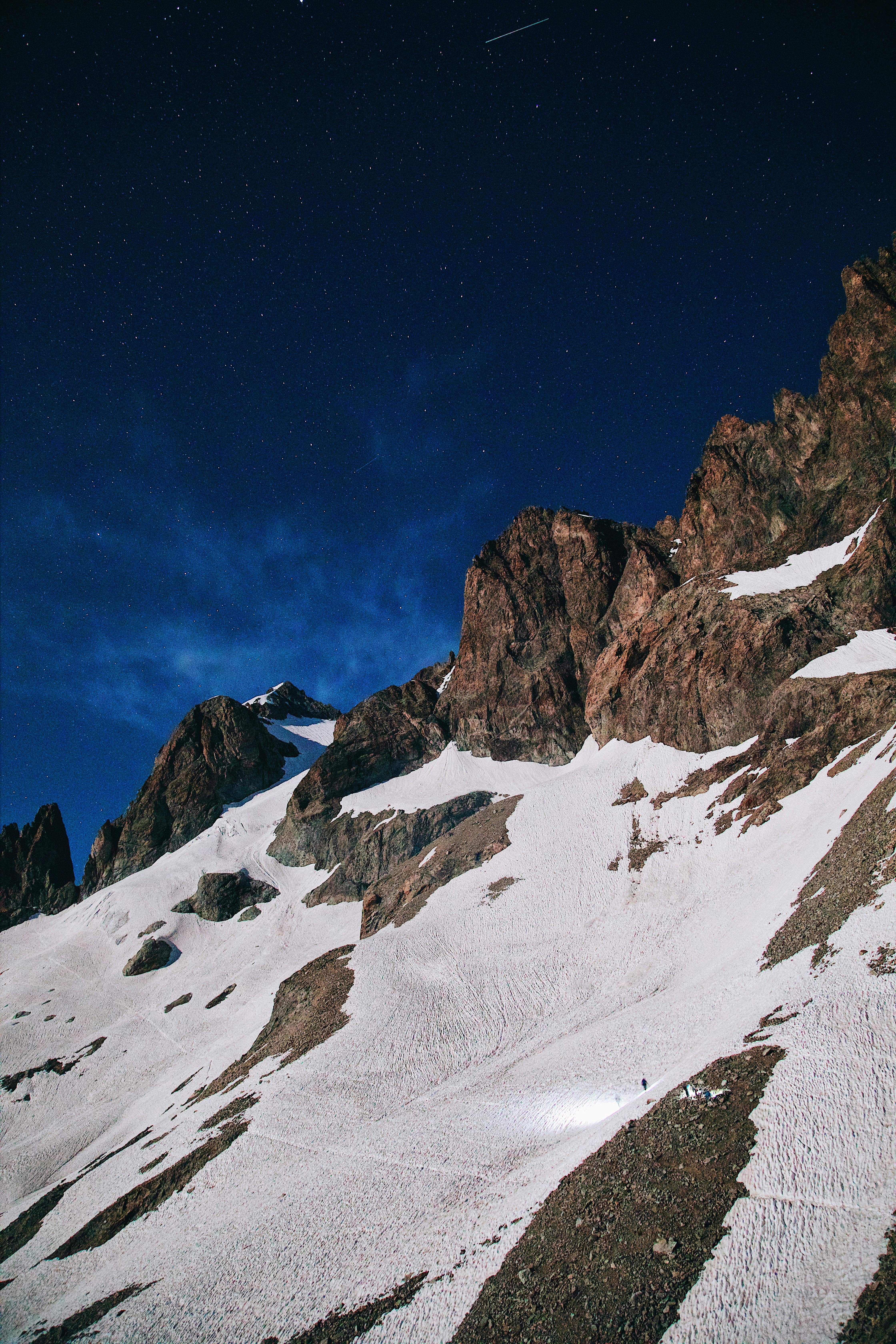 Stars illuminate the rugged snow-capped mountain slopes under a moonlit night sky.