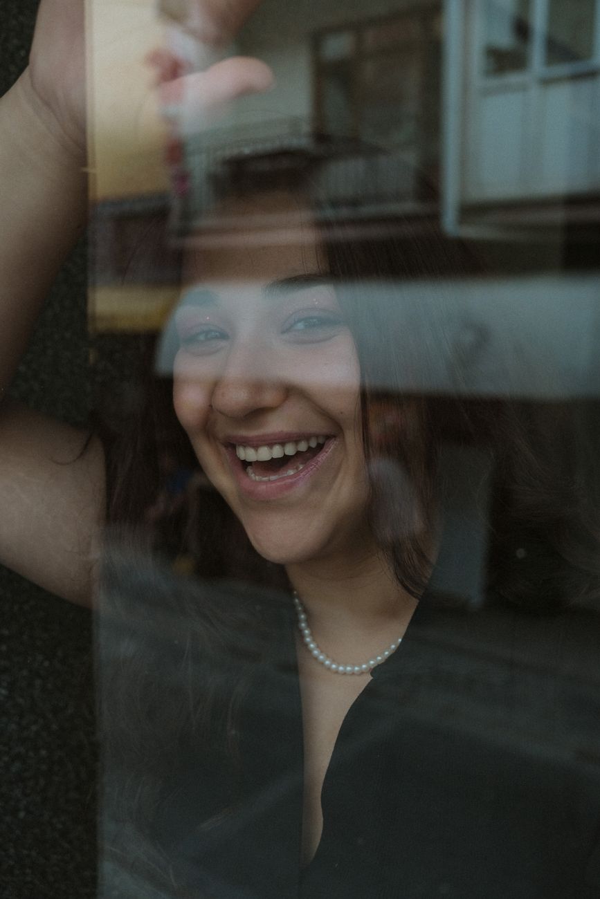 A joyful woman smiling behind a window, beautifully captured with reflections.